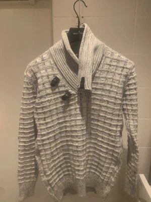 Grå stickad kofta från Battistini - Säljer en grå stickad kofta från Battistini med grovt rutmönster och bred krage. Koftan har snygga knappar och detaljer vid halsen, samt långa ärmar och ribbad nederkant. Perfekt för dig som gillar stilrena och mysiga plagg.