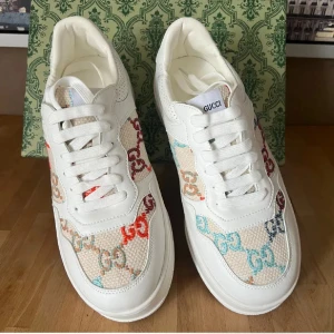 Gucci vita sneakers med färgglada GG - Gucci sneakers i vitt skinn och canvas med färgglada GG-loggor i rött, blått, beige och guld. Snygg chunky sula med präglad logga och guldfärgad GG-detalj på hälen. Klassisk snörning och rund tå. Perfekta för dig som vill sticka ut med lyxig streetstyle.