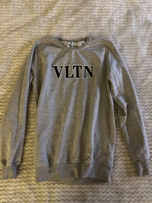 Grå sweatshirt från Valentino VLTN - Grå sweatshirt från Valentino med VLTN-tryck i svart och vitt på bröstet. Tröjan har rund hals, ribbade muddar vid ärmslut och nederkant samt långärmad passform. Mjuk och skön bomullskvalitet, perfekt för en chill och stilren look.