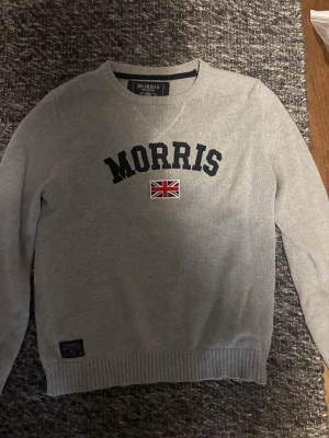 Grå stickad tröja Morris XL - Snygg grå stickad tröja från Morris i storlek XL med broderad logga och brittisk flagga på bröstet. Tröjan har rund hals, ribbade muddar och lång ärm. Perfekt för dig som gillar klassisk stil med en modern touch.
