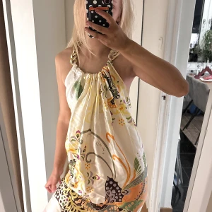 Blommig silkesklänning Amour de Pin'up - Unik kortklänning från Amour de Pin'up Paris i 100% silke. Klänningen har ett färgstarkt blommigt mönster i vitt, gult, grönt och orange, med snygga knytband vid axlarna. Luftig och flowy passform, perfekt för dig som gillar statement-plagg och boho vibes. Passar en s/m