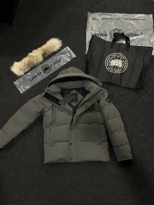 Mörkgrå pufferjacka från Canada Goose - Säljer en mörkgrå pufferjacka från Canada Goose med avtagbar päls på huvan. Jackan har snygga svarta detaljer, dragkedja och knappar framtill, samt logotyp på ärmen. Perfekt för kalla dagar med sin tjocka dunfyllning och stilrena look. Säljer då jag sparar till körkort😊