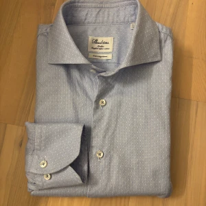 Blå slim fit skjorta från Stenströms - Snygg blå slim fit-skjorta från Stenströms med extra långa ärmar – perfekt för dig som gillar att kunna vika upp eller dra upp ärmarna utan att de känns för korta. Skjortan har ett diskret vitt prickmönster, klassisk krage och vita knappar. Tillverkad i mjuk och exklusiv bomull som ger en fräsch och stilren känsla. Nypris 2000 kr💸Mitt pris 199 kr.  Mått: Axelbredd 44 cm, armhåla 48 cm, ärmlängd 73 cm, längd 88 cm.