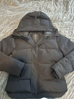 Svart pufferjacka från Canada Goose - Säljer en svart pufferjacka från Canada Goose i storlek L. Jackan har huva, dragkedja och knappar framtill samt en snygg broderad logotyp på armen. Den håller dig varm under kalla dagar. 