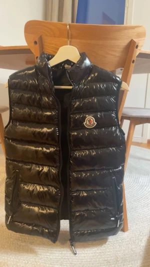 Svart dunväst från Moncler - Snygg svart dunväst från Moncler med glansig finish och klassisk logga på bröstet. Västen har ståkrage, horisontella quiltade paneler och dragkedja framtill samt två praktiska fickor med dragkedja. Perfekt för lager-på-lager och streetstyle.