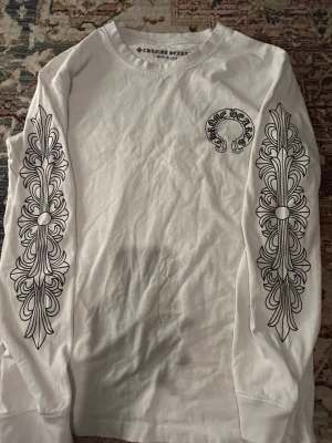 Vit långärmad t-shirt Chrome Hearts - Vit långärmad t-shirt från Chrome Hearts med svart tryck på ärmarna och bröstet. Snygga detaljer med grafiskt mönster längs båda ärmarna och logotyp på ryggen. Tillverkad i mjuk bomull och har en klassisk rund hals.