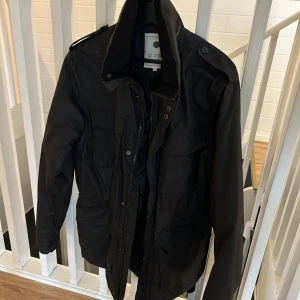 Svart fieldjacka - Svart fieldjacka med flera stora fickor framtill, hög krage och axelklaffar. Jackan har dragkedja och knappar, samt en stilren och funktionell design som passar perfekt till jeans. Materialet är slitstark polyester. Knappt använd.