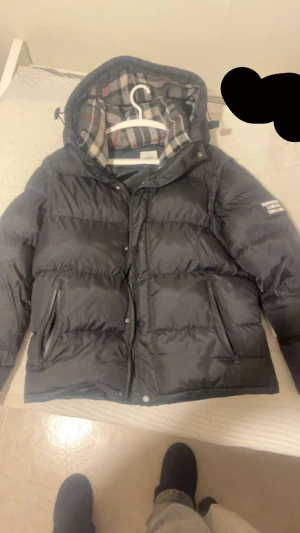 Svart pufferjacka Burberry med avtagbara ärmar - Svart pufferjacka från Burberry med avtagbara ärmar så du kan bära den som väst. Jackan har huva med klassiskt rutigt Burberry-foder, dragkedja och knappar framtill samt två sidofickor med dragkedja. Perfekt för dig som vill ha en flexibel och snygg jacka.