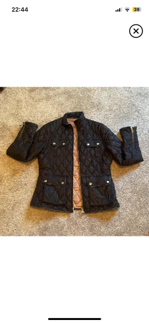 Svart quiltad jacka Barbour  - Quiltad svart jacka från Barbour Range Rover med snygga detaljer som silverfärgade knappar och dragkedja. Jackan har två stora fickor framtill, ståkrage och beige insida. Materialet är polyester och jackan har en normal passform med långärmade ärmar. Enda defekten kedjan/hängaren lite rostig