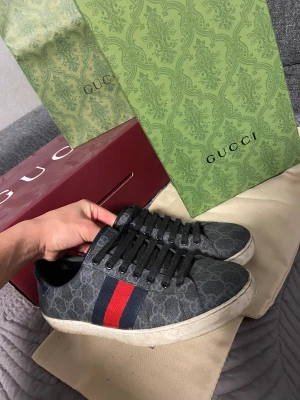 Gucci Ace - Snygga Gucci sneakers i mörkgrått med klassiskt GG-mönster och svarta snören. Sidan har en bred röd och marinblå rand, och hälen har metallicdetaljer i rött och blått. Yttermaterialet är canvas med skinn på insidan. Perfekt för dig som gillar lyxiga streetwear-vibes. Skorna är använda som ni ser. Hör av er vid funderingar. Pris kan diskuteras…
