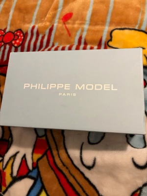 Philippe Model vita sneakers - Snygga vita Philippe Model sneakers med svarta detaljer och beige sula. Skorna har snörning och en rund tå, med en ikonisk sköld-logga på sidan. Ovandelen är i skinn och mocka, vilket ger en lyxig känsla. Perfekt för dig som gillar fransk streetstyle.  Sulan i skon är borta men man kan lägga in vilken Annan sula som helst använda ett tag 