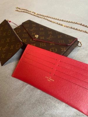 Louis Vuitton Monogram axelväska - Louis Vuitton axelväska med klassiskt monogrammönster i brunt och beige, guldfärgad kedja och matchande plånbok samt röd kortficka. Väskan har envelope-lock och detaljer i guld. Perfekt för dig som vill ha en ikonisk designeraccessoar med lyxig vibe.
