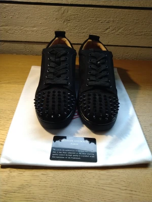 Christian Louboutin svarta sneakers med nitar - Svarta sneakers från Christian Louboutin med coola svarta nitar på tån och klassiska röda sulor. Skorna har snörning och är tillverkade i mocka med en rund tå. Insidan är beige och sulan har Louboutin-loggan i rött. Perfekt för dig som vill sticka ut.Det ni ser på bilderna ingår.