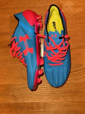 Blå och rosa Under Armour fotbollsskor stl 42 - Säljer ett par fotbollsskor från Under Armour i blått med rosa detaljer och snörning. Skorna har en rund tå och platt sula med dobbar för bra grepp på planen. Ovandelen har ett diskret hexagonmönster och insidan är gul. Perfekt för dig som vill sticka ut på matchen! Stl 42 men sitter mer som 41