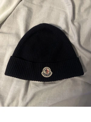 Svart/blå  Moncler mössa - Svart/blå  ribbstickad mössa från Moncler med klassisk logotyp framtill. Tillverkad i mjuk ull som håller dig varm och snygg. Perfekt accessoar för kyliga dagar och enkel att matcha med din jacka eller hoodie. Såklart Äkta!!