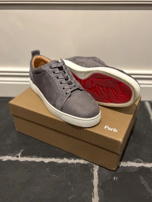 Grå mockasneakers Christian Louboutin - Snygga grå sneakers från Christian Louboutin i mocka med klassisk röd sula och vita kanter. Skorna har rund tå, snörning och lyxig känsla med läderfoder. Perfekta för dig som vill sticka ut med exklusiva detaljer och streetstyle-vibe.