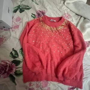 Rosa stickad tröja med paljetter - Säljer en fluffig rosa stickad tröja med guldiga paljetter över axlar och bröst. Tröjan har rund hals och långa ärmar med ribbade muddar. Perfekt för dig som vill sticka ut med lite extra glitter i vardagen.