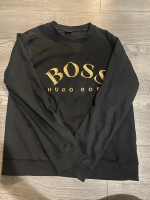 Svart sweatshirt från Hugo Boss - Svart sweatshirt från Hugo Boss med stort BOSS-tryck i guld på bröstet. Tröjan har rund hals, långa ärmar och ribbade muddar vid ärmslut och nederkant. Materialet är mjukt och känns som bomull, perfekt för en chill och stilren look.