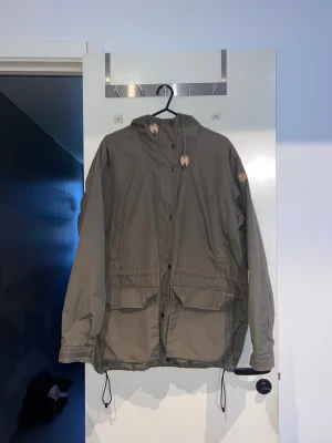 G-1000 klassisk modell från Fjällräven M  - Säljer en grön parkajacka från Fjällräven i storlek M. Jackan har rutigt fleecefoder i beige, grått och vitt, stora fickor framtill, dragsko i midjan och huva. Stängs med både dragkedja och knappar. Perfekt för kyliga dagar. OBS: Dragkedjan trasig se bild