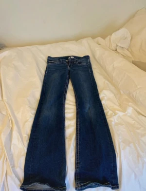 Blå bootcut jeans med vita sömmar - Snygga blå bootcut jeans med markerade vita sömmar och klassiska fickor både fram och bak, där bakfickorna har lock och knapp. Jeansen har låg midja och är tillverkade i jeansmaterial med dragkedja och knapp framtill.