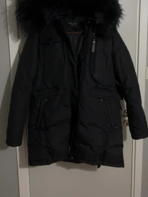 Hollies Livingo Long jacka - Svart lång dunjacka från Hollies med huva och äkta fluffig päls. Jackan har flera fickor, dragkedja framtill och snörning i midjan för snygg passform. Perfekt för kalla dagar och har en stilren look som funkar till det mesta. Har bara använts en vinter. 