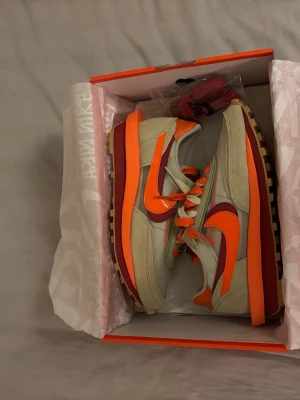 Nike LDWaffle S/C Blaze Orange sneakers - Nike LDWaffle S/C sneakers i beige mocka med orange och röda detaljer, dubbla Swoosh-loggor och platt gummisula med rutmönster. Skorna har snörning och cool retrostil, perfekt för dig som gillar färgstarka och unika sneakers.