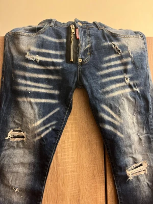 Blå skinny jeans med slitningar och dragkedja - Säljer ett par blå jeans med coola slitningar, blekta detaljer och en stor dragkedja framtill. Skinny fit och klassisk femficksmodell. Jeansen har en modern look med trasiga partier och vita färgstänk för extra edge.