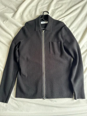 Samsøe Samsøe overshirt zip - Svart overshirt från Samsøe Samsøe med rak passform, klassisk krage och silverfärgad dragkedja framtill. Jackan har en enkel bröstficka och är tillverkad i ett grovt, ulligt material som ger en cool textur. Perfekt att slänga på över en t-shirt.