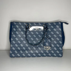 Guess Monogram Tote väska  - Snygg och rymlig Guess-väska i klassiskt monogrammönster. Perfekt som vardagsväska, skolväska eller jobbväska! Den har gott om plats för dator, böcker och allt du behöver under dagen. Enda deffekter är att nägra av prickarna på sidorna går av som ni kan se på bilderna men annars är den ny och i fint skick.  ✔ Skick: Mycket fint skick / Nära nyskick  ✔ Färg: Mörkblå / Navy ✔ Detaljer: Silverdetaljer, logoplatta fram, dubbla handtag + avtagbar axelrem ✔ Modell: Guess 4G Logo Tote ✔ Material: Slits