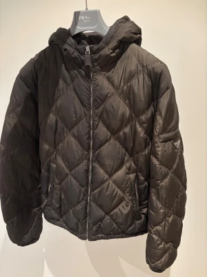 Prada pufferjacka - Quiltad mörkgrön pufferjacka från Prada med huva och dragkedja framtill. Jackan har ett snyggt rutmönster och ikonisk Prada-logga på ärmen. Materialet är glansigt och känns lyxigt, perfekt för dig som vill ha en varm och stilren jacka med exklusiv vibe. Passar large.