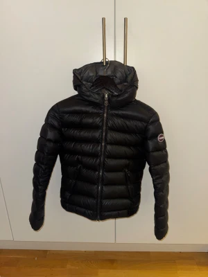 Svart dunjacka från Colmar - Säljer en svart dunjacka från Colmar med huva och dragkedja framtill. Jackan har ett stilrent, quiltat mönster och logotyp på ärmen. Fylld med naturligt dun för extra värme och har två fickor med dragkedja. Perfekt för kalla dagar och snygg till streetwear.