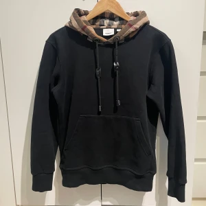 Svart Burberry hoodie med rutig huva - Svart hoodie från Burberry med klassisk rutig huva i beige, svart och rött. Tröjan har dragsko i huvan, magficka och ribbade muddar. Perfekt för dig som vill ha en stilren men ändå lyxig look. Pris går att diskuteras😁