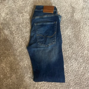 Blå jeans från Jack & Jones - Snygga blå jeans från Jack & Jones med klassisk femficksdesign och läderpatch bak i midjan. Jeansen har raka ben och slimfit passform för en cool look. Perfekta för dig som gillar en tidlös stil med lite edge.