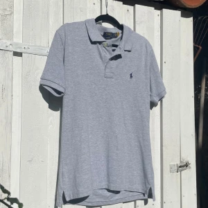 Grå pikétröja från Ralph Lauren - Klassisk grå pikétröja från Polo Ralph Lauren med korta ärmar, krage och två knappar framtill. Broderad marinblå logga på bröstet. Skön bomullskvalitet och stilren passform som ger en clean look.