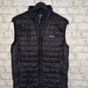 Svart quiltad dunväst från Patagonia - Svart dunväst från Patagonia med quiltad design och hög krage. Västen har dragkedja framtill och två sidofickor med dragkedja. Perfekt för lager-på-lager och outdoor. Diskret Patagonia-logga på bröstet. Snygg och funktionell för kyliga dagar.