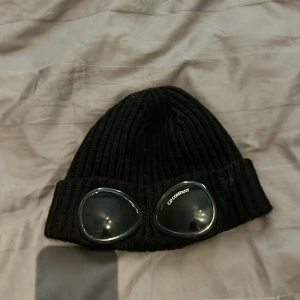 Svart C.P. Company goggle mössa - Svart ribbstickad mössa från C.P. Company med ikoniska goggle-detaljer framtill. Mössan är tillverkad i mjuk ull och har en uppvikt kant där de svarta glasögonlinserna sitter. Perfekt för dig som vill sticka ut med streetwear-vibe. Qr koden går att skanna och priset kan sänkas vid snabb affär!