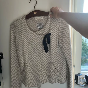 Beige kofta från Odd Molly med rosett - Superfin beige kofta från Odd Molly med zigzag-mönster och en söt mörkblå rosett framtill. Koftan har broderad logga i nacken, rund halsringning och är långärmad. Perfekt för lager-på-lager och chill dagar.