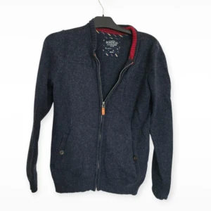 Mörkblå kofta från Bondelid med dragkedja - Mörkblå stickad kofta från Bondelid i storlek small. Koftan har dragkedja framtill, två fickor med knapp och snygga röda ribbade detaljer på insidan av kragen och ärmsluten. Perfekt för dig som gillar klassisk stil med en twist.