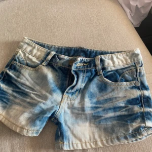 Blå tie-dye jeansshorts från Xando - Snygga blå jeansshorts från Xando med cool tie-dye effekt i ljusblått och vitt. Klassisk femficksmodell, låg midja och dragkedja med knapp. Perfekta för sommaren och ger en avslappnad vibe till din outfit.