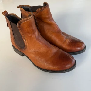 Bruna boots i skinn, strl 37 - Snygga bruna boots från Bugatti i äkta skinn. Skorna har elastiskt parti på utsidan och dragkedja på ”insidan”. Låg klack. Mjukt foder på insidan och robust sula för extra komfort. En liten reva vid tån på högerskon, se bild. 