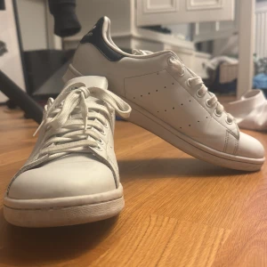 Adidas Stan Smith vita sneakers - Klassiska Adidas Stan Smith sneakers i vitt skinn med mörkblå detalj på hälen. Skorna har rund tå, snörning och diskreta perforerade ränder på sidorna. Perfekta för en clean och tidlös stil. Ikonisk Stan Smith-logga på plösen.