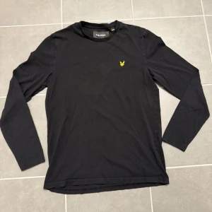 Svart långärmad tröja Lyle & Scott - Svart långärmad tröja från Lyle & Scott med klassisk rund hals och gul broderad logga på bröstet. Tillverkad i mjuk bomull som är skön mot huden. Perfekt för en clean och stilren look.