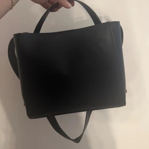 Svart axelväska från Gina Tricot - Stilren svart axelväska i slät skinnimitation från Gina Tricot. Väskan har en fyrkantig form, två handtag och ett rymligt huvudfack med innerficka som stängs med dragkedja. Perfekt för dig som gillar minimalistisk och modern design.