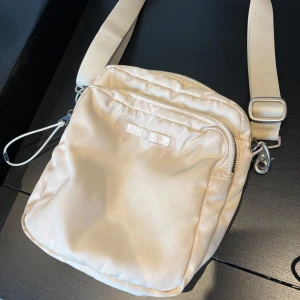 Beige axelväska från Our Legacy - Snygg beige axelväska från Our Legacy med justerbar rem och praktiska dragkedjor. Väskan har en stilren, fyrkantig form och är tillverkad i ett mjukt, glansigt syntetmaterial. Perfekt för dig som vill ha en clean och minimalistisk look.