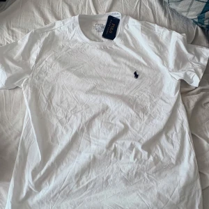 Vit t-shirt från Polo Ralph Lauren - Vit t-shirt från Polo Ralph Lauren i bomull med klassisk rund hals och broderad marinblå logga på bröstet. Modellen är slim fit och har korta ärmar. Perfekt basic-plagg med stilren design och skön passform.