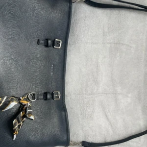 Marinblå  axelväska från Puccini i skinn - Stilren marinblå axelväska från Puccini i präglat skinn med silverfärgade spännen och dragkedja. Väskan har en dekorativ scarf, flera fack inuti och ett mönstrat innerfoder i beige. Perfekt för dig som vill ha en klassisk och praktisk väska med snygga detaljer.