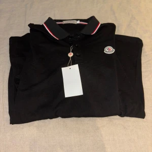 Svart pikétröja från Moncler M - Svart pikétröja från Moncler med klassisk krage och knäppning framtill. Kragen har snygga röda och vita ränder och tröjan pryds av Monclers ikoniska logga på bröstet. Tillverkad i bomull för en skön och stilren känsla.