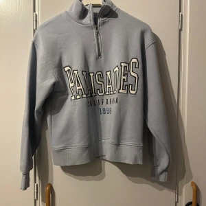 Ljusblå half zip sweatshirt Gina Tricot - Ljusblå sweatshirt från Gina Tricot med broderad text 'Palisades California 1998' framtill. Tröjan har half zip, hög krage och långa ärmar. Mjuk insida och relaxed passform, perfekt för chill dagar.