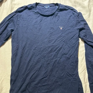 Mörkblå långärmad tröja från GANT - Snygg mörkblå långärmad tröja från GANT med rund halsringning och diskret logga broderad på bröstet. Tillverkad i mjuk bomull som känns skön mot huden. Perfekt för dig som gillar stilrena och klassiska plagg.