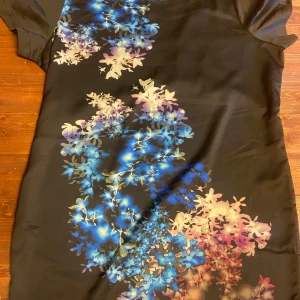 Svart omlottklänning med blommigt mönster - Snygg svart omlottklänning i glansigt material med korta ärmar. Framsidan har ett färgstarkt blommigt mönster i blått, lila och rosa toner. Klänningen har knytband i midjan och en mjuk, följsam känsla.